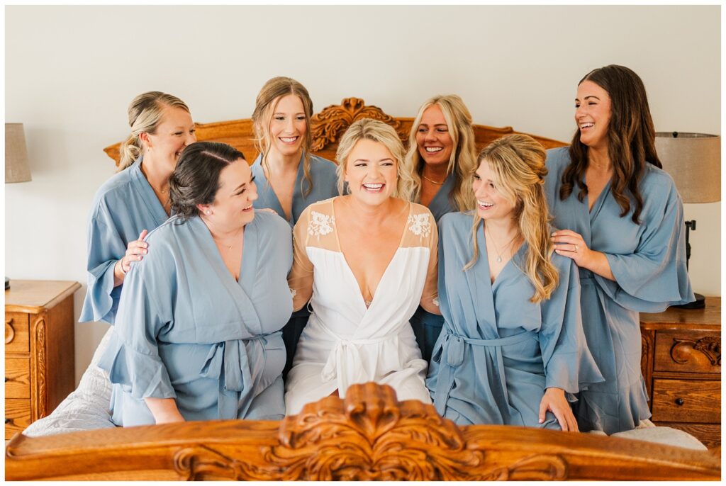 bride laughing with bridesmaids while wearing robes and sitting on a bed
