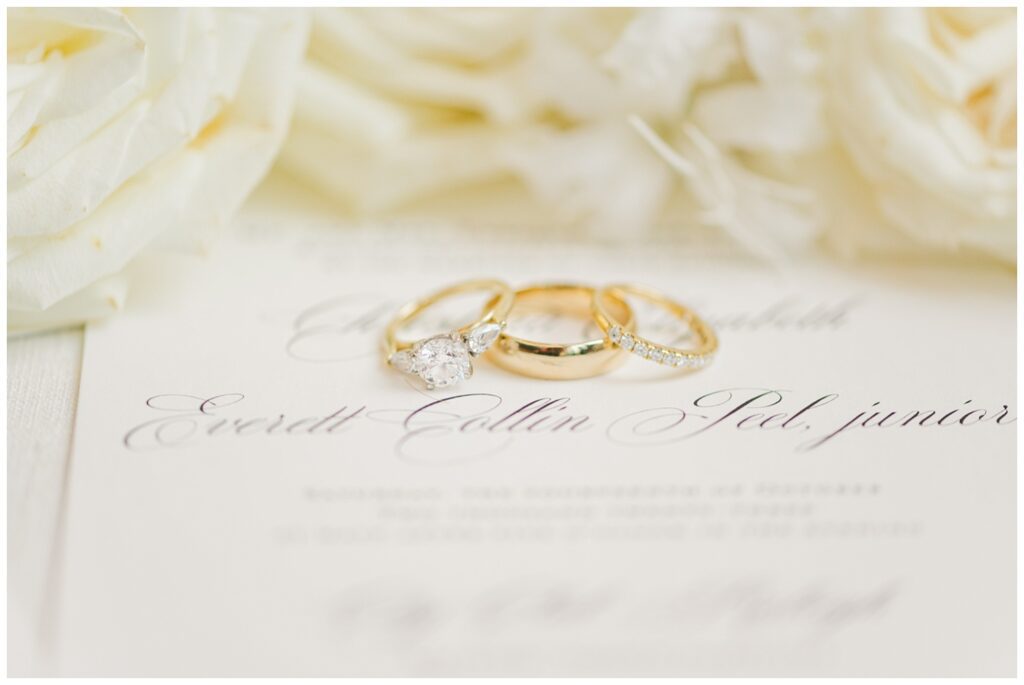 gold and diamond rings stacked on top of wedding invitation in front of white flowers