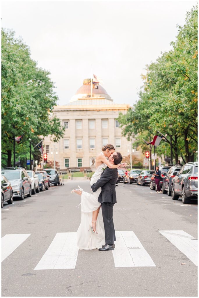 groom picking up bride for a kiss in the crosswalk in front of City Hall in Raleigh