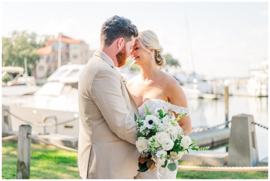 groom holding the bride's waist while she holds her bouquet next to the boats 