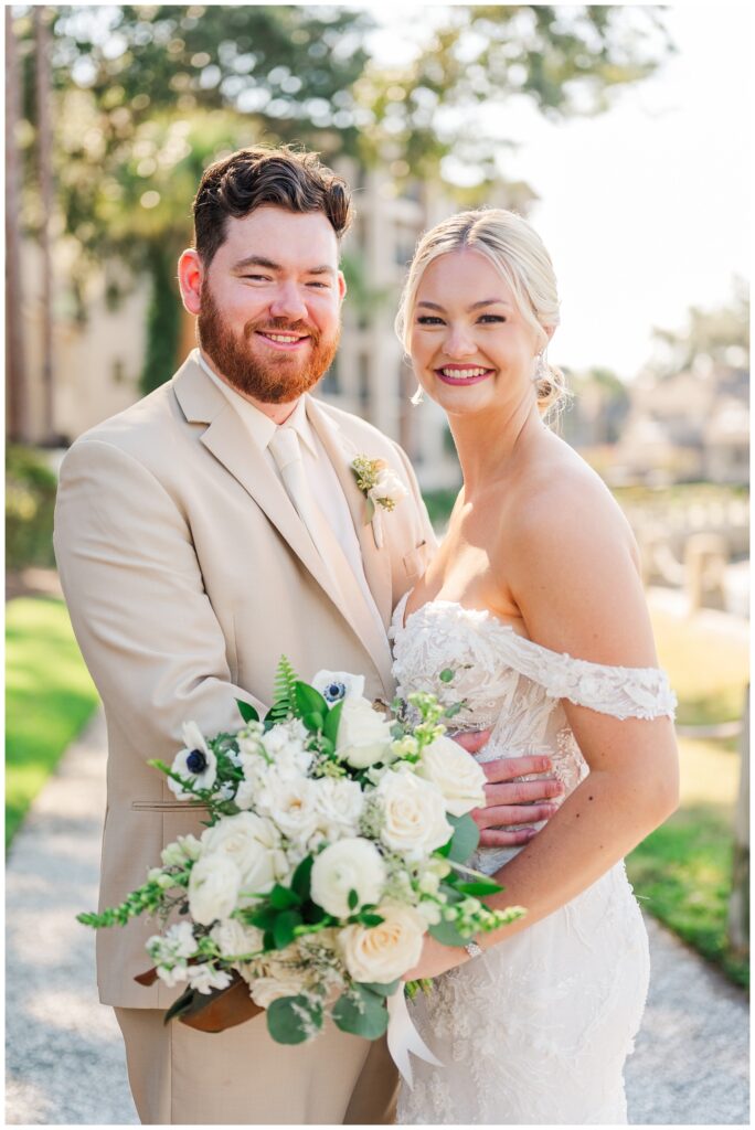 groom holding the bride's waist while she holds her bouquet next to the water