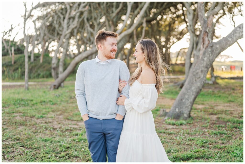 engagement couple smiling together in front of live oaks at Fort Fisher