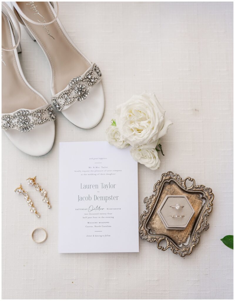 white wedding invitation on a flat lay mat with bride's shoes, white flowers, and jewelry