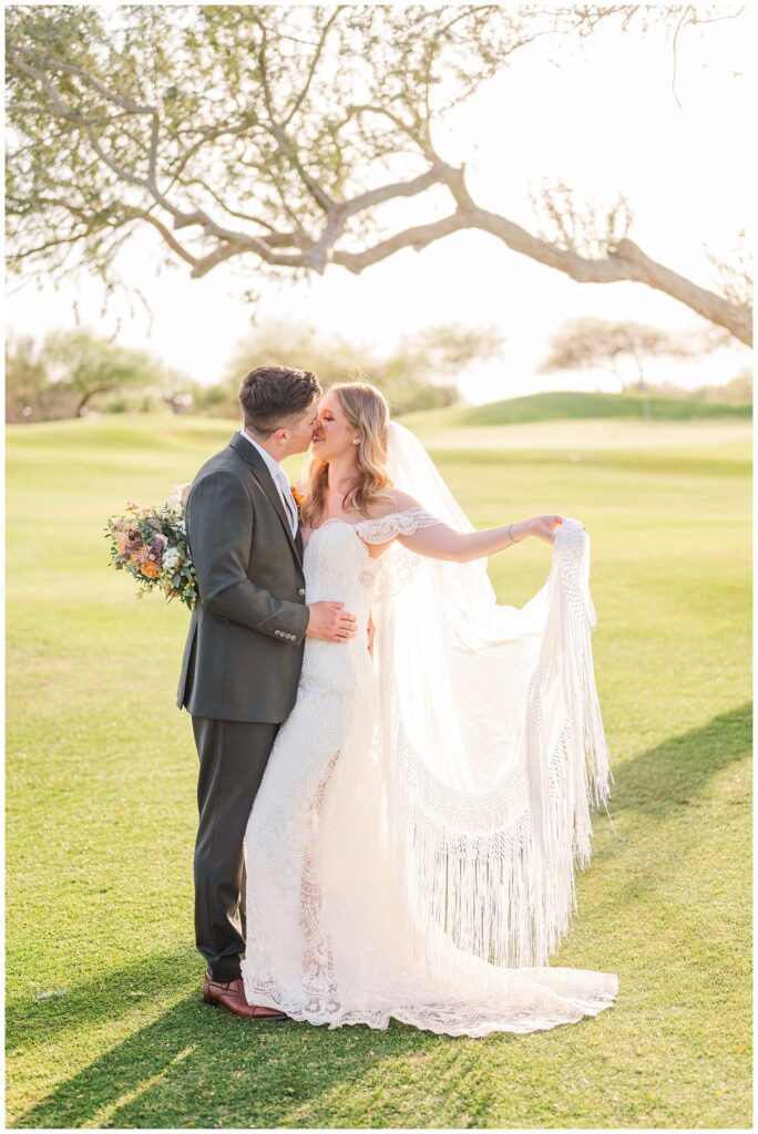 groom kissing the bride while she is holding her veil on the golf course