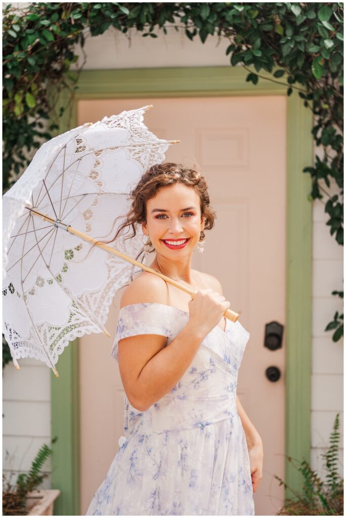 bride holding a white lace parasol in front of a pink and green door outside 