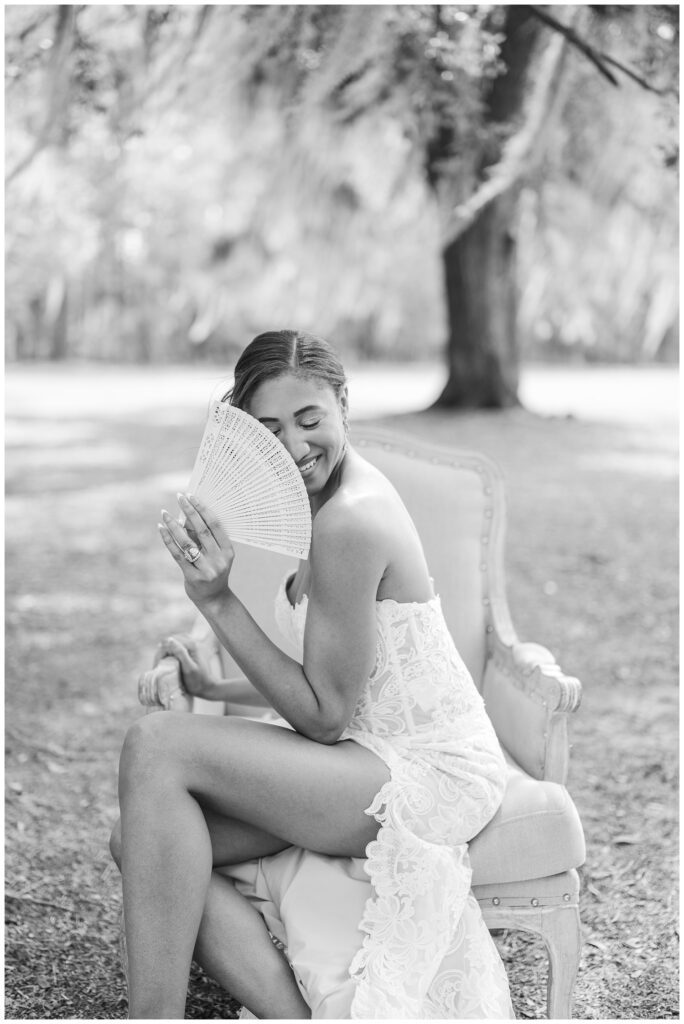 bride sitting in a wingback chair covering part of her face with a fan at Hewitt Oaks Venue