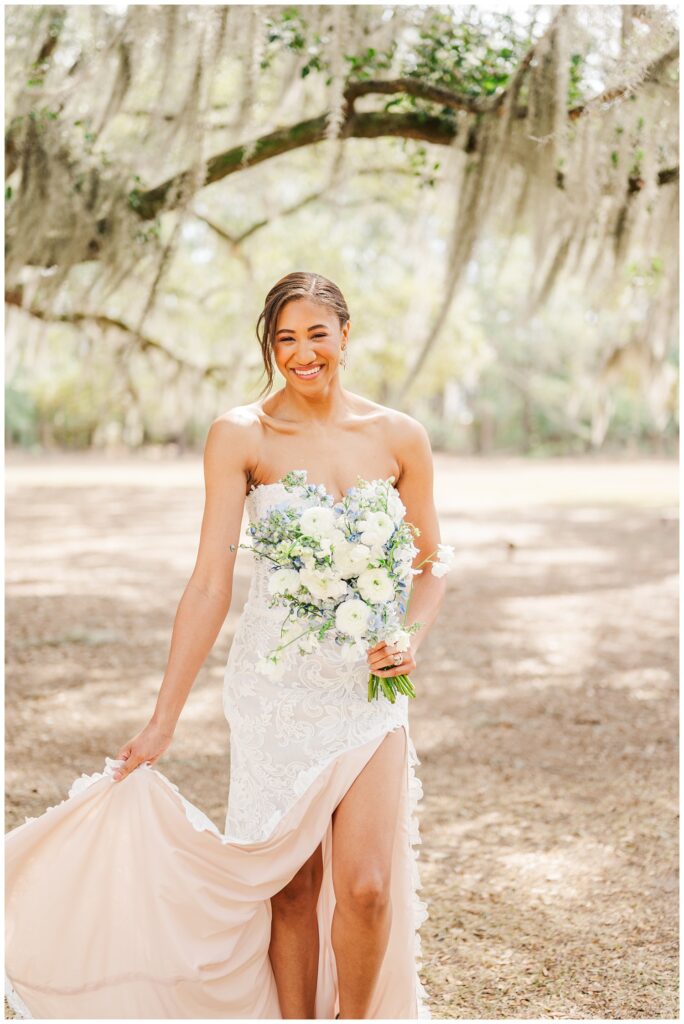 bride holding the train of her strapless lace wedding dress and walking with a blue and white bouquet