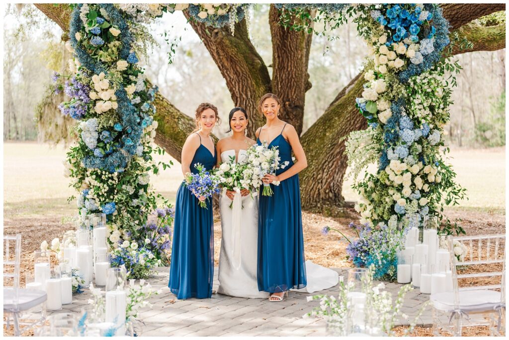 bride posing with her bridesmaids under a live oak tree surrounded by large blue and white flowers on an arch