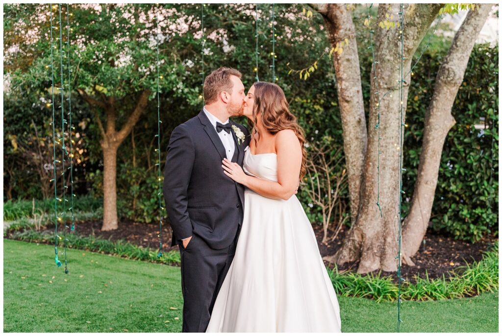 bride and groom share a kiss in front of string lights outside during cocktail hour