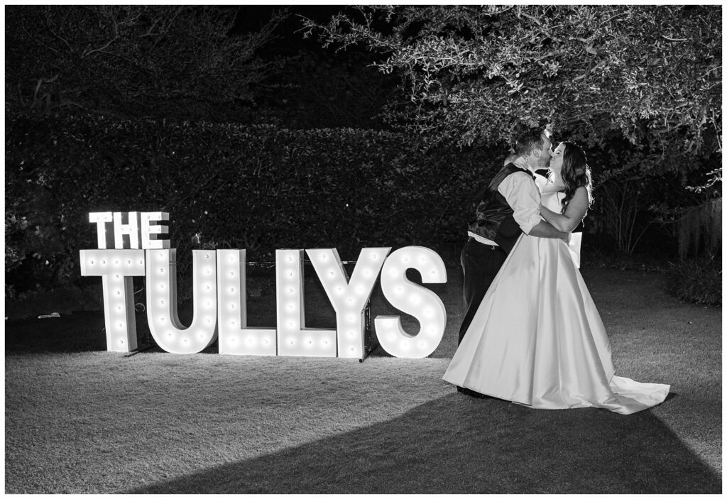 bride and groom share a kiss at night next to their lighted letters sign outside